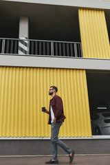 Good walk. Side view of handsome young bearded man in casual wear and eyeglasses holding a disposable cup while walking through the city street