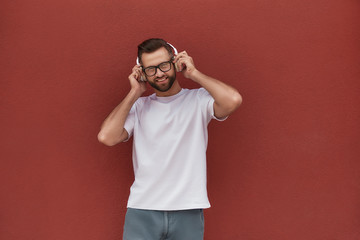 Enjoying my favourite song. Portrait of handsome and cheerful man in eyeglasses wearing headphones and listening to the music while standing against red wall outdoors