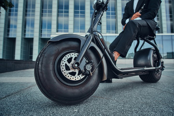 Elegant businessman in black suit is sitting on his electrical scooter near his office.