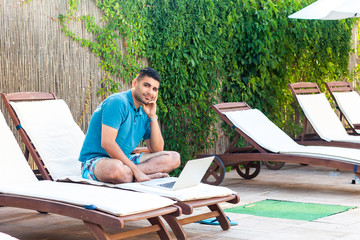 Portrait of happy handsome bearded young adult freelancer man in blue t-shirt and shorts sitting on deckchair with laptop on poolside and looking at camera. Lifestyle concept, outdoor, summer vacation