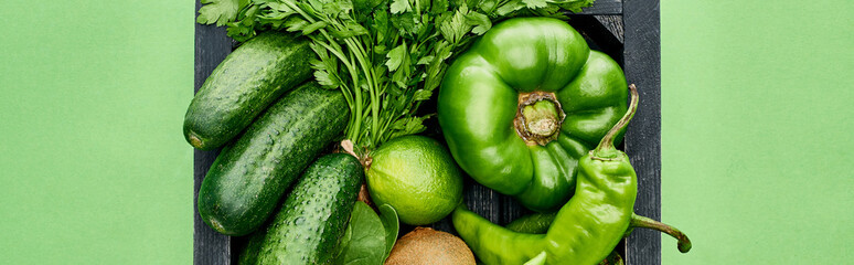 panoramic shot of fresh cucumbers, kiwi, limes, peppers and greenery in wooden box