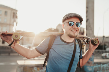 Fototapeta premium Happy hipster guy posing with skateboard in sunset in summer evening. Portrait of handsome stylish man model in blue shirt, sunglasses and hat on city street background