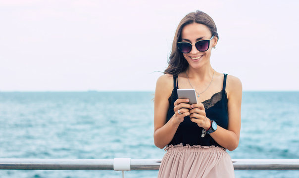 Vacation, travel, relaxation, freedom and happy time. Beautiful smiling young brunette woman in sunglasses is using her smart phone on the sea background