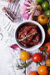Sun dried tomatoes in a white ceramic bowl surrounded by cherry tomatoes, garlic, spices and herbs on wooden table