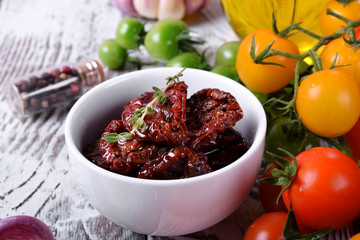 Sun dried tomatoes in a white ceramic bowl surrounded by cherry tomatoes, garlic, spices and herbs on wooden table