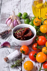 Sun dried tomatoes in a white ceramic bowl surrounded by cherry tomatoes, garlic, spices and herbs on wooden table