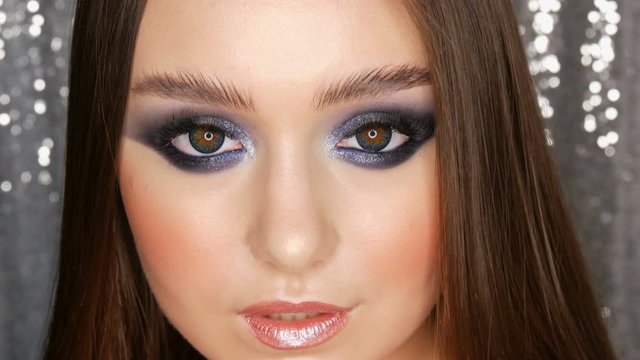 Beautiful sexy young girl with evening blue make-up smoky eyes long hair and cosmetic colored contact lenses posing in model studio on the silver background.