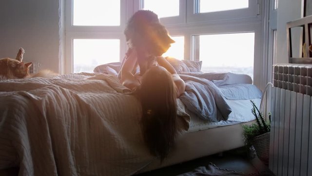 Beautiful romantic young brunette woman lying on bed by the window with her lovely cat during amazing sunset
