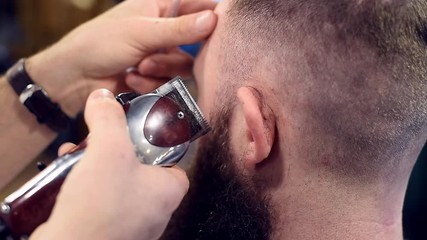 Close up filming of using special brush and shaving hair with electric razor machine near ear on male head with back view. Short haircut for bearded man. Cropped view of barber hands in action