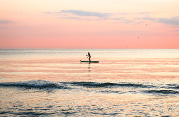 Fototapeta premium Woman's silhouette on a paddle board (SUP) at a beautiful pink pastel gradient sunset.