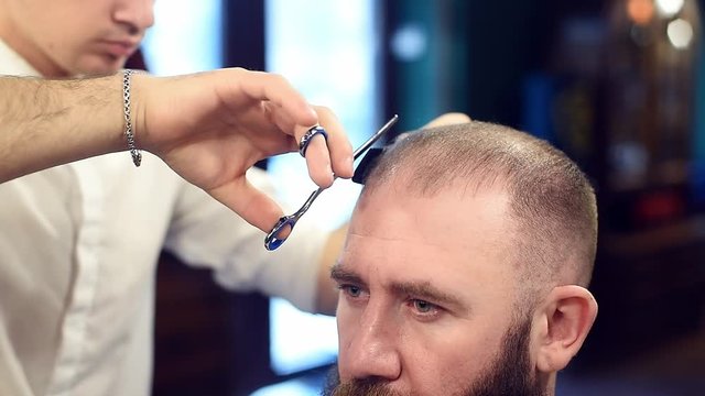 Cutting short hair with scissors and comb helping. Male hairstyle. Cropped view of male barber and man brunette bearded client during hair cutting process in hair salon barbershop. Blurred background