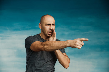 A young bald man in a gray T-shirt on a blue background, angrily screams and points a finger.
