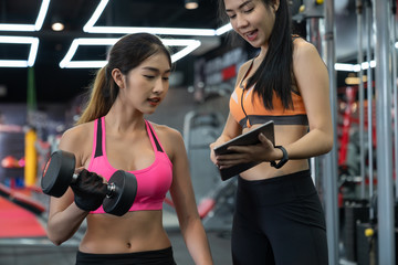 Asian woman lifting a dumbbell with assistance of her personal trainer at public gym.