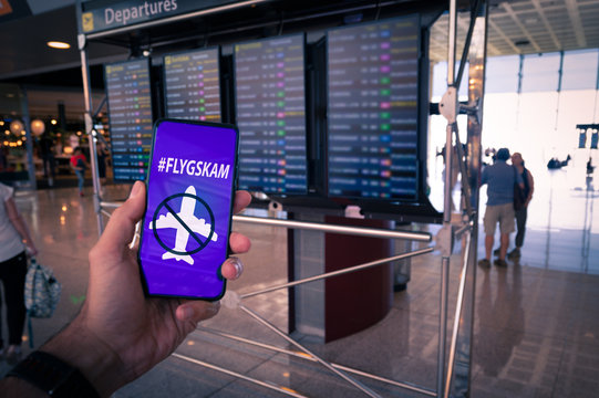 Hand holding a smartphone with Flyskam message on screen with airport timetables as background. Flygskam or flight shame in Swedish refers to the feeling of being ashamed or embarrassed to board a