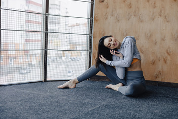 Young attractive fitness girl sitting on the floor near the window on the background of a wooden wall, resting on yoga classes and talking on phone