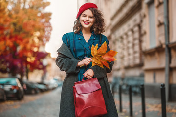 Outdoor autumn portrait of young happy smiling lady wearing checkered dress, coat, beret, holding red leather bag, orange fallen leaves, posing in street of European city. Copy, empty space