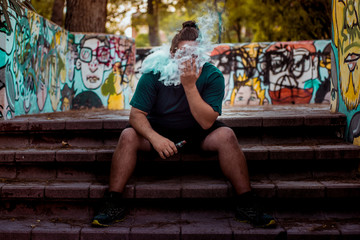 Young man with long hair and big beard sitting on the stairs, smoking in a park with graffiti.