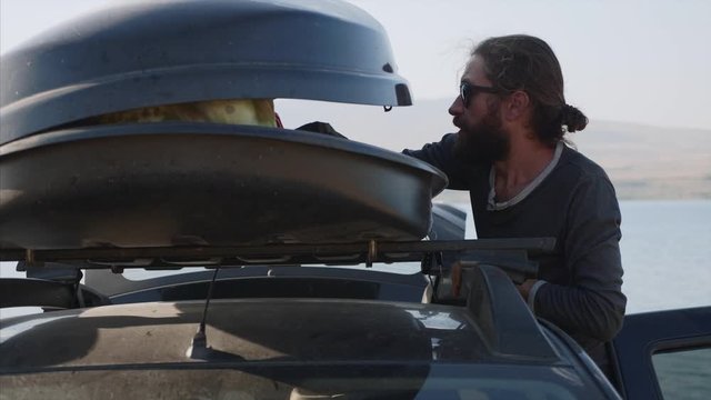 Young bearded man taking out a camping equipment from car roof box and throwing it on the ground, side view. Traveler is came to the lake and preparing for camping.