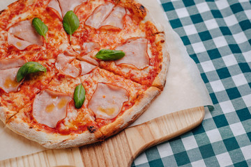 Neapolitan pizza with ham, cheese, arugula, basil, tomatoes sprinkled with cheese on a wooden board on a tablecloth in a cell with a place for text