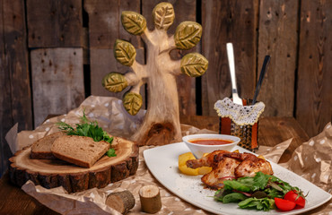 chicken with potatoes, tomatoes and herbs, a Cup of sauce on a white plate and pieces of black bread on a tray of a bar of wood