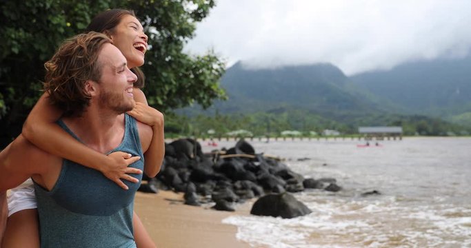 Two young people playing having fun laughing in love. Interracial couple piggybacking, boyfriend carrying Asian girlfriend doing piggyback on hawaiian beach in Hanalei Bay, Kauai, Hawaii travel.