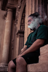 Obraz na płótnie Canvas Young man with a beard uses an electronic cigarette sitting on a historic building.