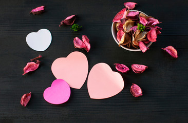 Dry air petals of pink roses lie on the table in a white Cup, next to them are pink  paper sheets in the form of hearts for messages on a black background