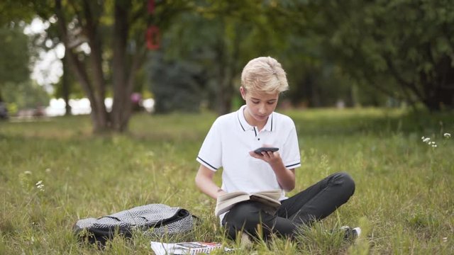Boy studying in park, reading book and being distracted by phone