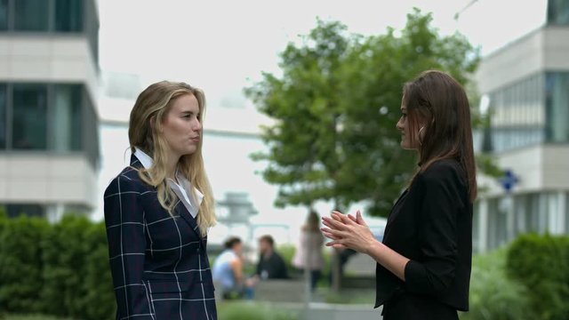 Standing outdoors, in a modern business campus, two women have a discussion
