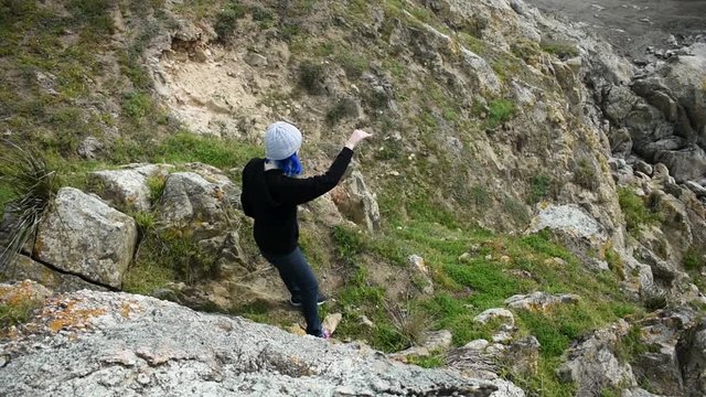 Blue haired girl with a wool cap throwing a rock down a cliff towards the sea
