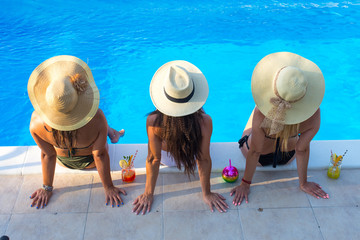 three beautiful young woman with sun hat sitting by the poolside of a resort swimming pool