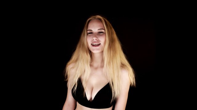 Lit from below, a busty blonde woman looks directly at the camera, laughing and shrugging suggestively