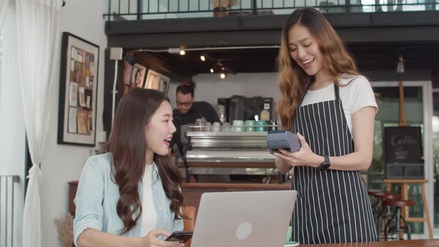 Young Asian freelance women pay contactless at coffee shop. Asian happy girl barista waiter wear gray apron holding credit card reader machine for customer using mobile phone scan pay in cafe.