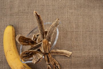 Long form banana chips in a glass bowl on a gray burlap, top view