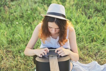 Girl teenager in hat playing guitar sitting on the grass