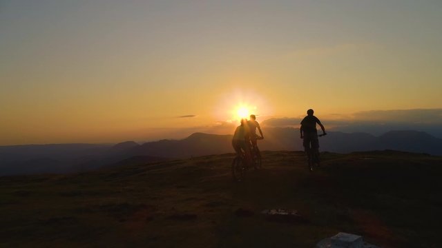 AERIAL, SILHOUETTE, SUN FLARE: Three mountain bikers ride up to end of a grassy hill at sunrise and high five after a successful bicycle trip. Friends celebrate an awesome downhill biking adventure.