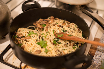 Pasta noodles with mushrooms and herbs cooked in a pan with a wooden spatula on a dirty gas stove in the kitchen, cooking, dinner, parsley