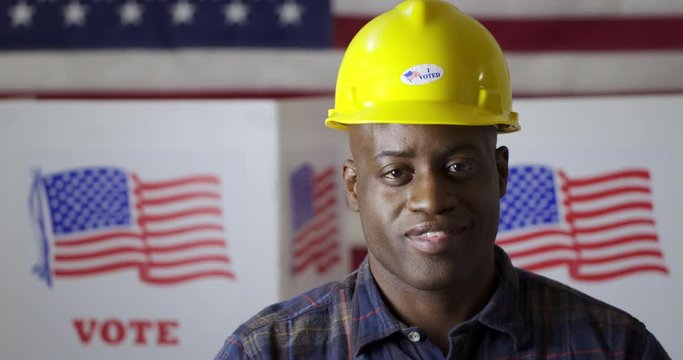 Close up African American man in plaid shirt puts on hard hat with "I Voted" sticker while facing camera and poses, smiling in front of polling booths with US flag. Large US flag behind