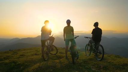 SUN FLARE: Three unrecognizable friends observe the evening nature from a mountain top after a fun mountain bike journey in Slovenia. Mountain cyclists watch the sunrise before a fun downhill race. - Powered by Adobe