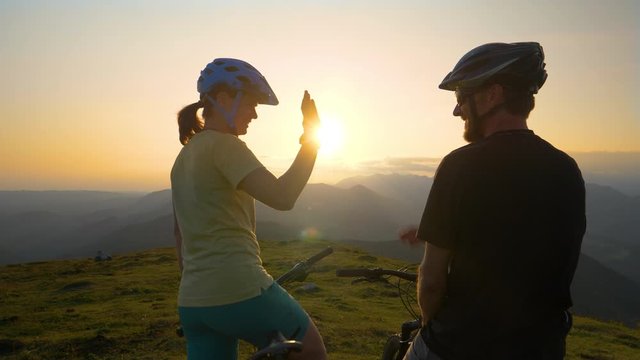 CLOSE UP LENS FLARE SLOW MOTION: Happy man and woman high five over the sunset after a fun mountain biking trip in the picturesque mountains. Friends celebrate an awesome cross country cycling journey