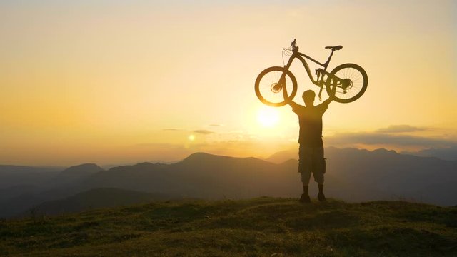 SILHOUETTE, LENS FLARE, SLOW MOTION, COPY SPACE: Cheerful athletic man holds his mountain bicycle above his head after a fun cross country trip in the picturesque golden lit Slovenian mountains.