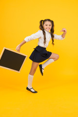 Fototapeta na wymiar Starting school with a smile. Happy small child going to school on yellow background. Active little girl in school uniform holding blackboard. Back to school season, copy space