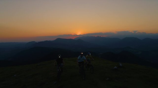 AERIAL: Flying in front of a group of mountain bikers pedalling their bikes up a steep grassy hill at night. Three friends wearing headlamps go on a cross country bicycle trip early in the morning.
