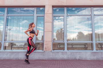 Fototapeta na wymiar A beautiful girl in summer runs in the city on a run, the background is a building with large glass windows, an active lifestyle, free space for text, sportswear leggings top and sneakers