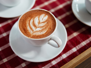 A cup of fresh brewed cappuccino coffee with tree branch flower pattern in a white mug served on a red white checkered tray in cozy city cafe background