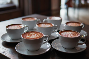 Set of freshly brewed cappuccino cups with milk foam latte art served on bartend table in cozy city cafe lit with natural window light