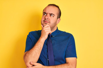 Young businessman wearing denim casual shirt and tie over isolated yellow background with hand on chin thinking about question, pensive expression. Smiling with thoughtful face. Doubt concept.