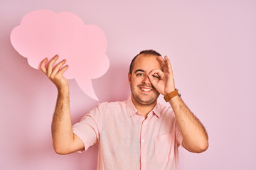 Young man holding cloud speech bubble standing over isolated pink background with happy face smiling doing ok sign with hand on eye looking through fingers