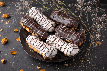 Eclairs or profiteroles with chocolate and custard cream on a metal plate on a dark background. Traditional French dessert. Empty space for design text template. Top view.