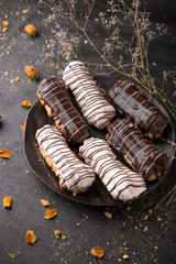 Eclairs or profiteroles with chocolate and custard cream on a metal plate on a dark background. Traditional French dessert. Empty space for design text template. Top view.